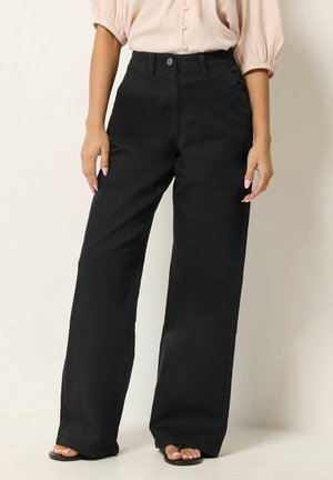 Mujer que lleva pantalones negros de pierna ancha y talle alto con bolsillos delanteros, combinados con una blusa beige y sandalias negras de punta abierta.