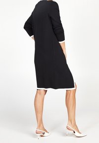 Černé pletené šaty s dlouhými rukávy, bílými manžetami a lemem. Mají boční rozparky a jednoduchý, pohodlný střih. Kombinovány s bílými lodičkami na zadním pásku.