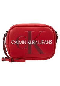 Červená obdélníková crossbody taška s nastavitelným popruhem, na přední straně s velkým embosovaným logem "CK" a bílým logem "Calvin Klein Jeans".
