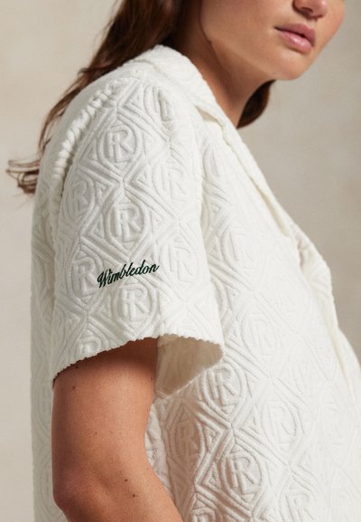 Femeie care poartă un tricou alb texturat cu mânecă scurtă, cu „Wimbledon” brodat pe mânecă.