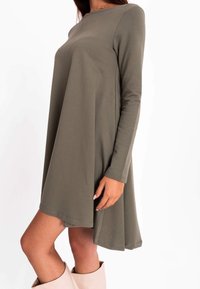 Olivgrünes langärmeliges Kleid, A-Linien-Silhouette, weicher Stoff, runder Ausschnitt, Länge bis zur Mitte des Oberschenkels, abgesteppten Säumen, kombiniert mit hellen Stiefeln.