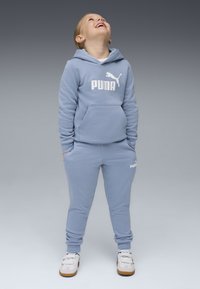 Sweatshirt azul-claro e calças de fato combinando com um logo Puma branco. O conjunto inclui um capuz, bolso frontal e punhos canelados. Os sapatos são brancos com solas castanhas.