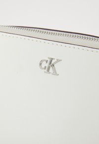 Baltas odinis rankinukas su sidabriniu užtrauktuku ir iškilniu "CK" logotipu priekyje. Lygi tekstūra su siuvinėjimo detalėmis aplink kraštus.