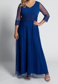 Long blue dress featuring a V-neck, sheer sleeves, embroidered waist, and flowing skirt. Sparkling accents enhance the bodice design.