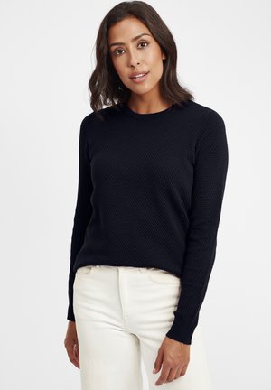 Marineblå teksturert strikkegenser med rund hals og lange ermer, kombinert med lyse jeans. Genseren har en figurnær, uformell stil.