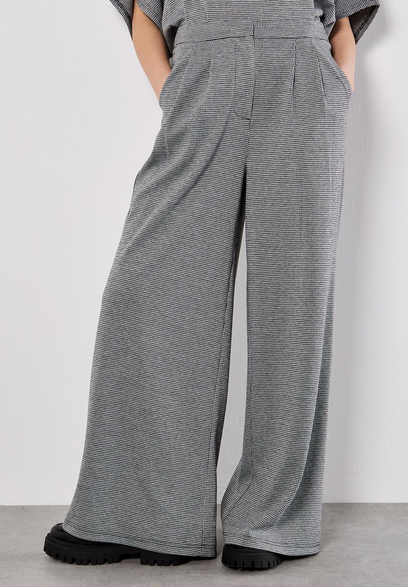 Breda byxor i ett svart och vitt rutmönster. Tillverkade av ett texturerat material med sidofickor och hög midja, matchat med kraftiga svarta skor.
