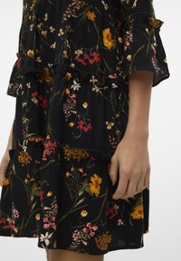 Vestido preto floral com camadas em babados, apresentando flores amarelas, vermelhas e brancas. Feito de um tecido leve com uma textura suave.