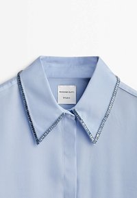 Chemise bleu clair à col avec une texture lisse. Le col est orné d'une bordure décorative en perles. L'étiquette indique "MASSIMO DUTTI STUDIO".