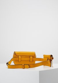 Piccolo marsupio in pelle giallo senape con tracolla regolabile e mini pochette incorporata, su un piedistallo bianco contro uno sfondo bianco.
