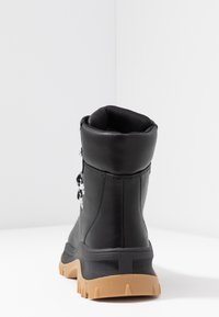Botín de cuero negro con un cuello acolchado, suela de goma texturizada y detalles de cordones. Presenta un acabado liso y una forma de punta redondeada.