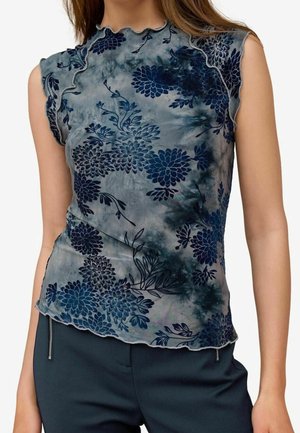 Femme portant un haut sans manches bleu-gris avec des motifs floraux en velours bleu foncé et un ourlet ondulé, associé à un pantalon foncé avec des détails zippés.