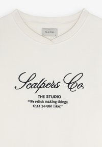 Crème T-shirt met korte mouwen en een klassieke ronde hals. Zwarte geborduurde tekst leest "Scalpers Co. THE STUDIO" en een citaat daaronder.