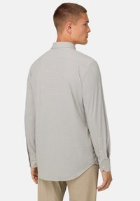 Maglietta grigia chiara a maniche lunghe con un motivo sottile, colletto a bottoni e polsini a un bottone, vista dal retro, infilata in pantaloni beige.