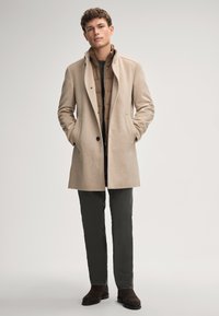 Beige wollen blend overcoat met een hoge kraag, knoopsluiting aan de voorkant en zijzakken, gelaagd over een bruine pufferjack, gecombineerd met donkere broeken en bruin laarzen.