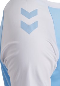 Kortærmet sportsjakke med hvide og lyseblå sektioner. Fremhæver markante blå Chevron-mønstre på skulderen, glat tekstur.