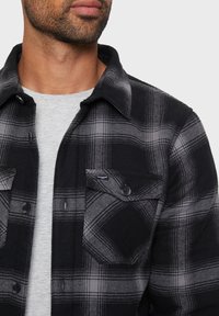 Mann trägt ein schwarz-graues kariertes Hemd über einem hellgrauen Rundhalsausschnitt-T-Shirt, mit Fokus auf dem Oberkörper und dem Hals.
