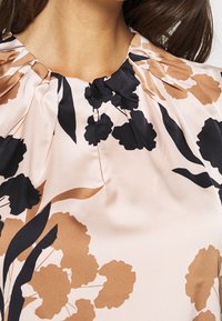 Seidenbluse mit floralem Design, die schwarze und braune Blüten auf einem hellrosa Hintergrund zeigt. Faltiger Ausschnitt mit einem gerafften Detail.