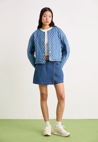 Τζάκετ τζην με ανοιχτό μπλε floral σχέδιο, κοντή σχεδίαση; συνδυασμένο με φούστα τζην σε μπλε απόχρωση και μπεζ αθλητικά παπούτσια με λευκές κάλτσες.