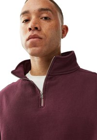 Maroon zip-up huppari korkean kauluksen kanssa, jossa on hopeinen vetskari ja pehmeä kangas, päällä valkoinen t-paita.