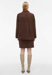 Veste en daim marron avec une coupe sur mesure, associée à une jupe en daim marron assortie. Texture lisse, design simple, portée avec des bottes beiges jusqu'aux genoux.
