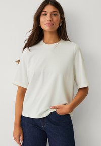 T-shirt beige clair à manches courtes avec un col rond et une coupe décontractée, associé à un jean en denim foncé. Texture lisse, design minimaliste.