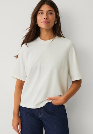 T-shirt beige clair à manches courtes avec un col rond et une coupe décontractée, associé à un jean en denim foncé. Texture lisse, design minimaliste.