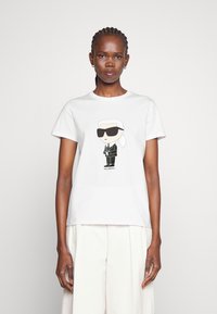 Balta kokvilnas T-krekliņš ar multfilmu attēlu, kurā attēlots tēls saulesbrillēs un melnā uzvalkā, centrā drukāts. Īsas piedurknes, apaļa kakla izgriezums.