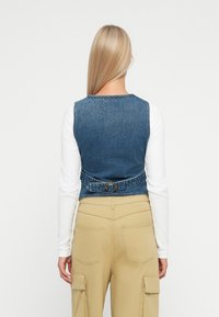 Μπουφάν από τζιν με μπλε πλύσιμο, λεπτομέρεια με πόρπη στο πίσω μέρος και εφαρμοστή σχεδίαση. Συνδυάζεται με λευκή μακρυμάνικη μπλούζα και καφέ cargo παντελόνια.