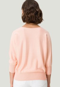 Femme aux cheveux bruns ondulés mi-longs, portant un pull léger pêche à manches longues et un pantalon blanc, de dos par rapport à l'appareil photo.