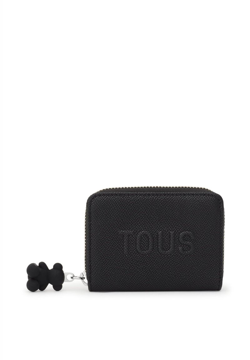 Cartera de cuero negro texturizado con el logotipo "TOUS" en relieve, cierre de cremallera y un charm en forma de oso negro adjunto. Diseño compacto y rectangular.