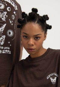 Young woman with bantu knot hairstyle wears dark brown t-shirt with white mystical butterfly and sun graphic, neutral background.