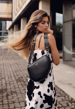 Schwarze Leder-Crossbody-Tasche mit einer gewölbten Form, silberner Hardware und verstellbarem Tragegurt. Getragen über einem weißen Kleid mit schwarzen Punkten.