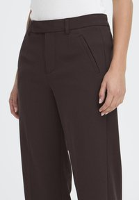 Pantalons ajustés marron avec une taille moyenne, des poches avant et une texture lisse, montrés de la taille vers le bas, avec une main reposant sur la jambe.