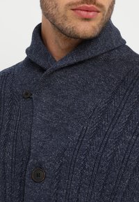 Marinblå stickad cardigan med sjalkrage, prydd med kabelstickat mönster och två mörka knappar. Mjuk textur, medel tjocklek.