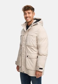 Giacca isolante beige con cappuccio, grandi tasche frontali, chiusura frontale con zip e bottoni a pressione, caratterizzata da dettagli neri e una texture liscia.