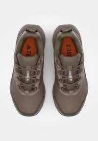 Para brązowych butów do biegania terenowego Adidas Terrex z sznurowadłami, siateczkową tkaniną i pomarańczowymi wkładkami, widziana z góry na białym tle.