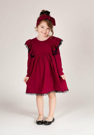 Bordowa sukienka z długim rękawem z falbanami na ramionach i koronkową lamówką u dołu, zestawiona z czarnymi balerinami i pasującą opaską na głowę.