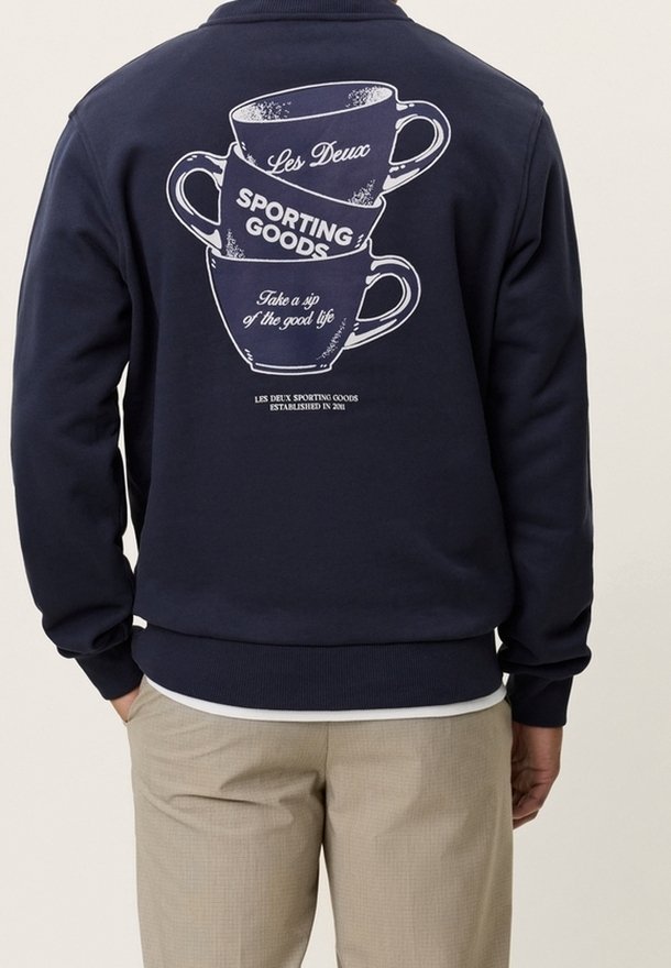 Personne portant un sweat-shirt bleu marine avec un motif blanc représentant trois tasses de café empilées et du texte dans le dos.