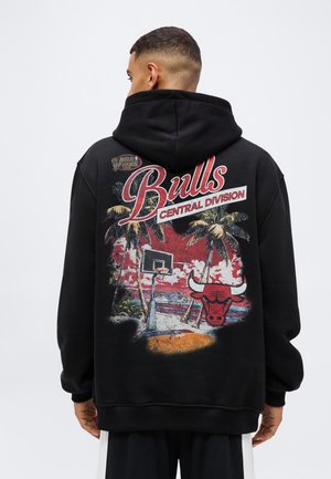 Homme portant un sweat à capuche noir avec le logo des Chicago Bulls, un panier de basket, des palmiers et le texte "Central Division" au dos.