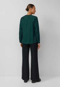 Ciemnozielona koszula z długim rękawem, okrągłym dekoltem i plisowanym tyłem, w zestawieniu z czarnymi spodniami z szerokimi nogawkami o wysokim stanie oraz czarnym obuwiem.