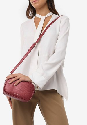 Rød glitrende crossbody taske med en tekstureret overflade, flad form og en lang justerbar skulderrem, holdt mod en lys bluse og brune bukser.