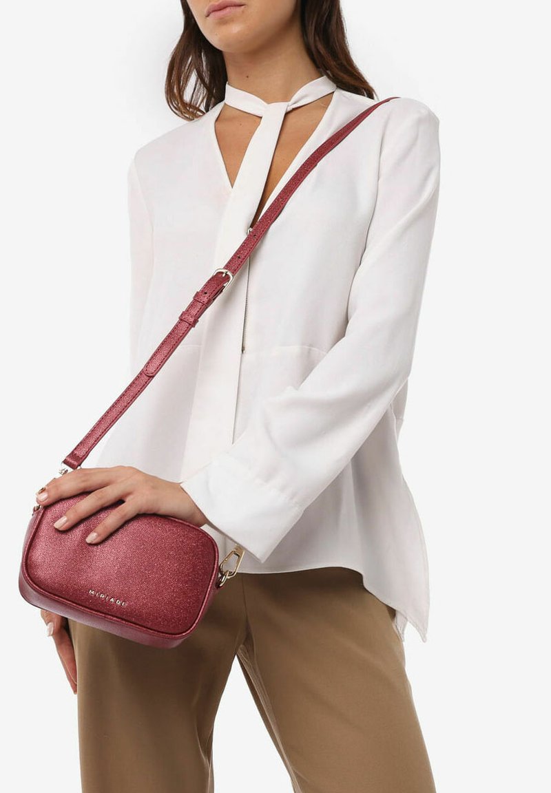 Bolso cruzado rojo con purpurina, con una superficie texturada, forma plana y una correa larga y ajustable, sujetado contra una blusa clara y pantalones beige.