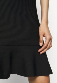 Un vestito nero a costine presenta un orlo svasato, un materiale texturizzato e un design aderente. Una mano è posizionata sulla zona della vita.