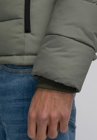 Chaqueta acolchada de un verde oliva claro con cremallera negra y puños de punto verdes. Tejido texturizado con un acabado suave que se muestra de forma destacada.