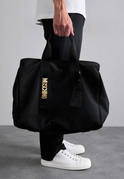 Juodas nailono rankinys su dviem rankenomis, auksiniu "MOSCHINO" logotipu, tekstūriniu akcentu ir lagaminų etikete. Paprastas fonas, kasdinis apranga.