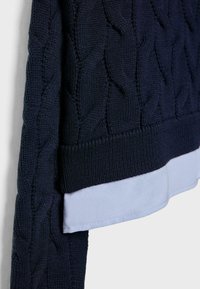 Maglione blu navy a maglia grossa con orlo e polsini a coste, indossato sopra una camicia azzurro chiaro. La texture è spessa e intrecciata con un motivo in rilievo.
