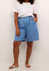 Shorts in lino azzurro chiaro con trama, con elastico in vita, tasche laterali e vestibilità ampia, abbinati a una t-shirt bianca e sandali.