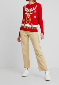 Punainen neulottu villapaita, jossa on poron kuva sarvineen, ruskea kasvot ja lumihiutaleita. Yhdistetty beigenvärisiin housuihin ja valkoisiin lenkkareihin.