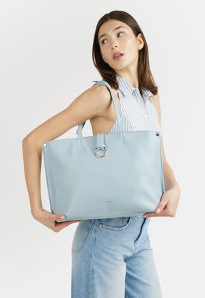 Donna con maglietta a righe senza maniche e jeans che tiene una grande borsa a spalla azzurra con dettaglio ad anello su sfondo bianco.