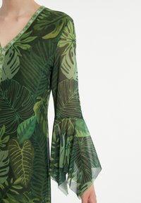 Grünes durchsichtiges Kleid mit V-Ausschnitt, ausgestelltem Ärmeln und einem Blattmuster in verschiedenen Grüntönen. Elegante Textur und Designdetails.